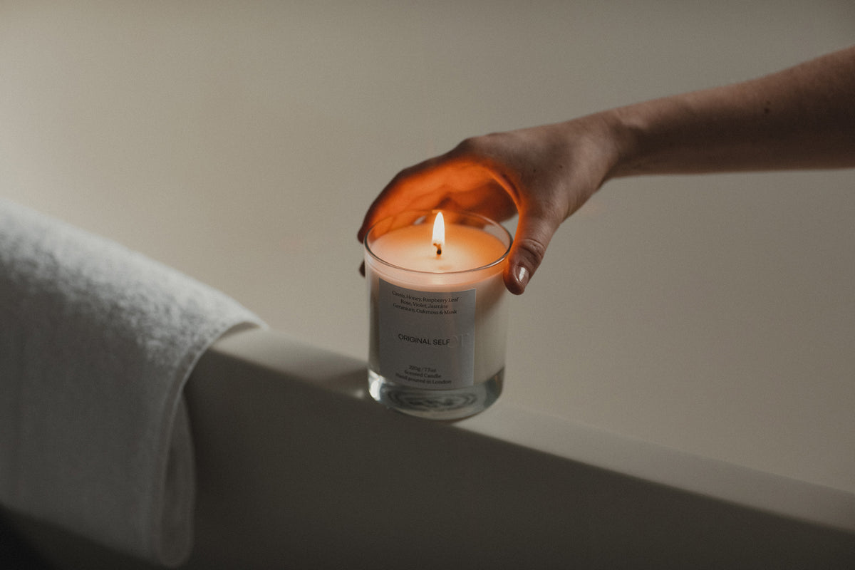 A hand holding an Original Self candle on the side of a bath tub.