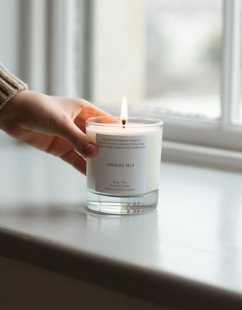 A hand placing  a lit Original Self candle on a windowsill.